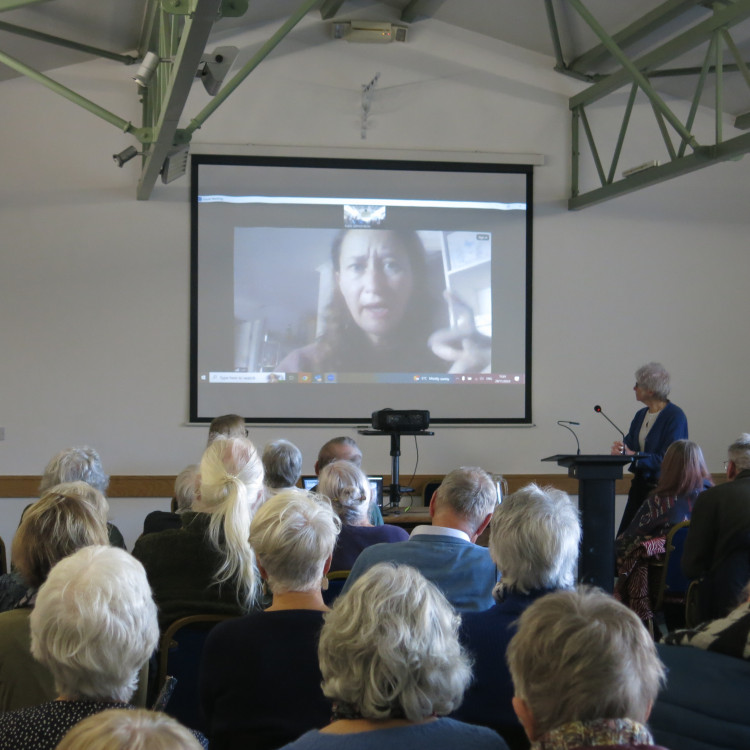 CAPTION: Ruth James (Oxfam International Regional Humanitarian Co-ordinator for the Middle East and North Africa) on zoom, with Jenny Williams at the lectern (chair of Mid Somerset Oxfam Group).
