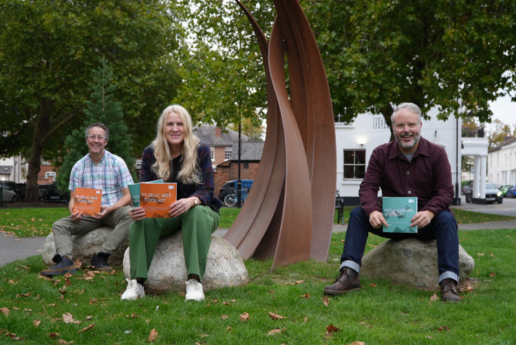  Paul Roberts (Arts Manager); Cllr. Ella Billiald; and  Johnathan Branson (Projects & Development Manager, Arts) proudly sharing  Warwick District Council's new Arts and Cultural Strategy and Public Art Toolkit,  in front of 'Down by the River’, a piece of public art outside the Royal Spa  Centre, Leamington, by Spencer Jenkins