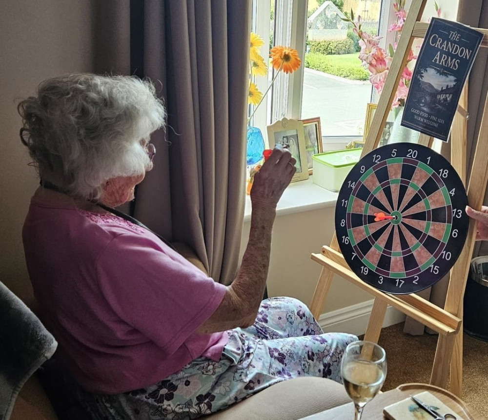 Crandon Springs resident Margaret enjoying a game of darts and a glass of wine while relaxing in her room during the pop-up pub event.